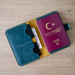Tüm bölmeleri dolu şekilde ahşap zeminde üstten fotoğraflanan Camel – Petrol Noir & Co. Lyra hakiki pueblo deri pasaport cüzdanı.